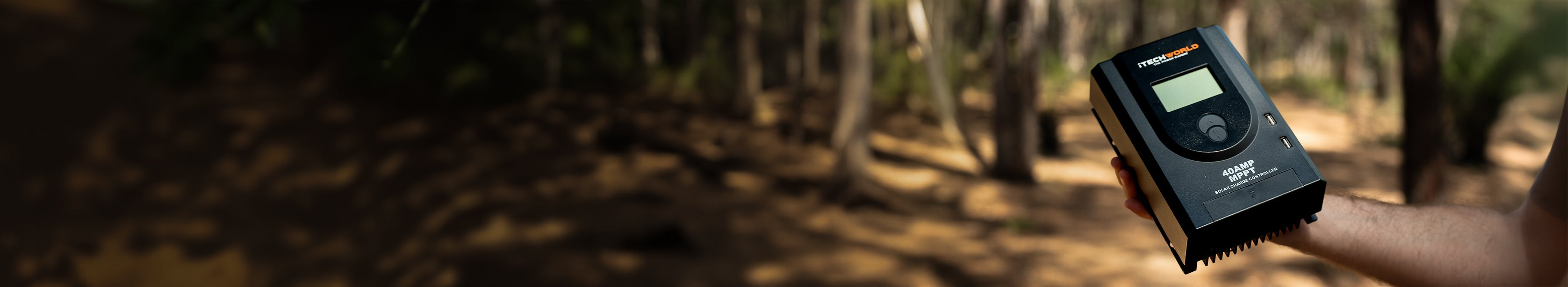 A person holds a black electronic device with a screen and buttons, standing outdoors in a sunlit forest with trees and shadows in the background.