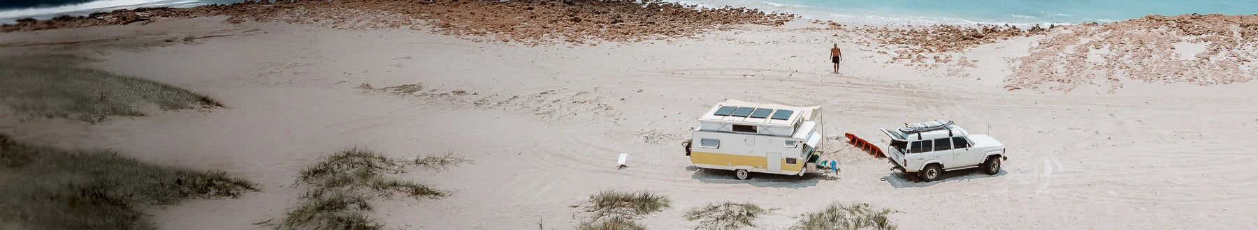A white SUV towing a yellow and white camper is parked on a sandy beach near the ocean, with a person standing by the water’s edge.