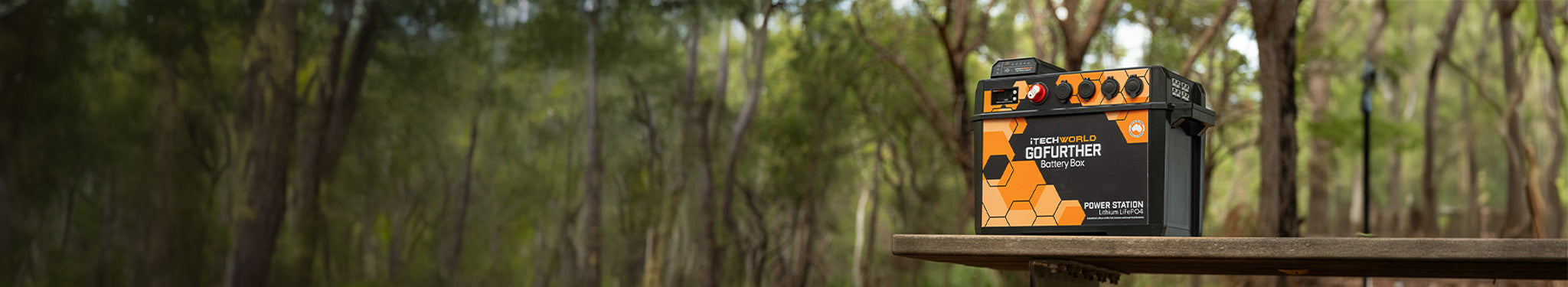 An orange and black portable power station sits on a wooden surface outdoors, surrounded by a forest with tall, blurred trees in the background.