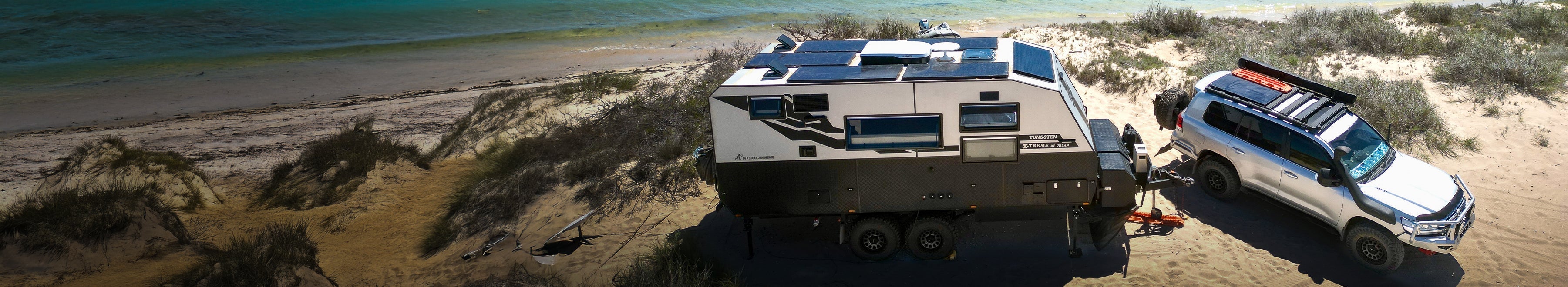 A white off-road vehicle tows a large, modern camper trailer with solar panels, parked on sandy terrain beside a blue coastline and sparse vegetation.