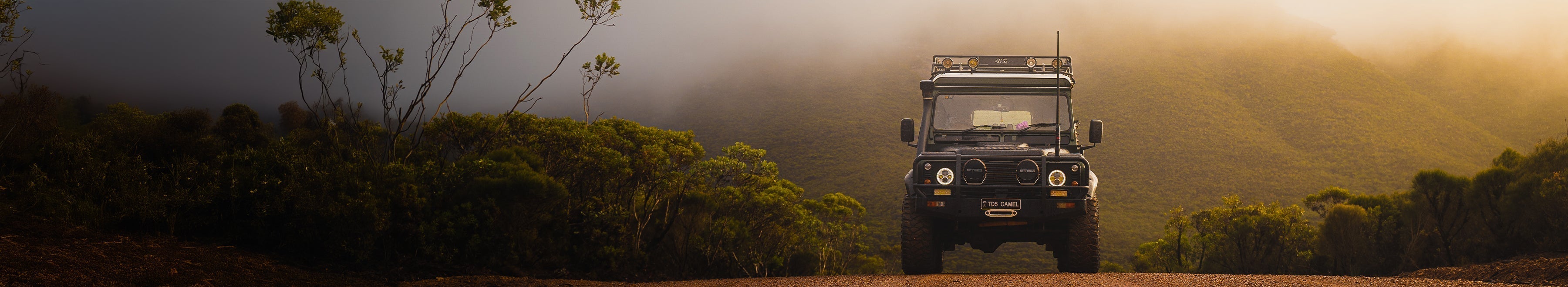 A black off-road vehicle drives on a dirt road surrounded by green bushes, with misty mountains and a golden sky in the background.