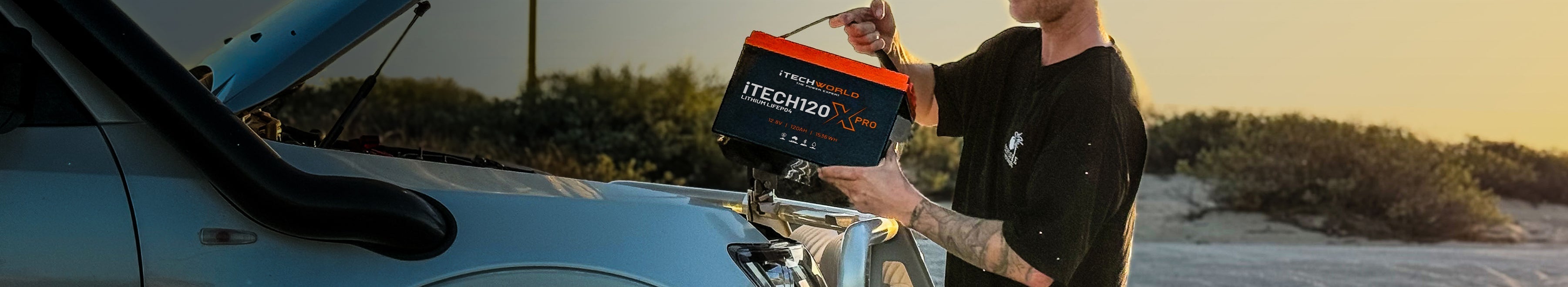 A person holds a large battery labeled “TECH120” while installing it into the engine compartment of a white vehicle outdoors at sunset.