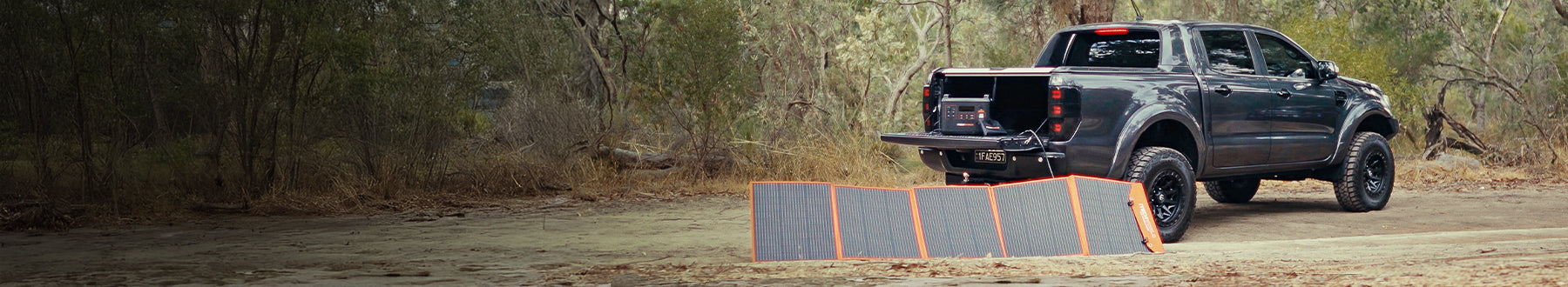 A black pickup truck is parked in a wooded area with portable solar panels set up on the ground behind it, connected to the truck’s tailgate.