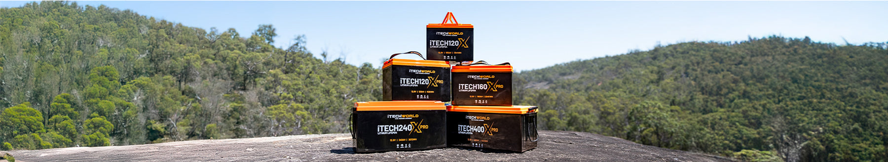 Five orange and black Trekoolah lithium batteries are stacked on a large rock outdoors, with green trees and hills in the background under a clear sky.