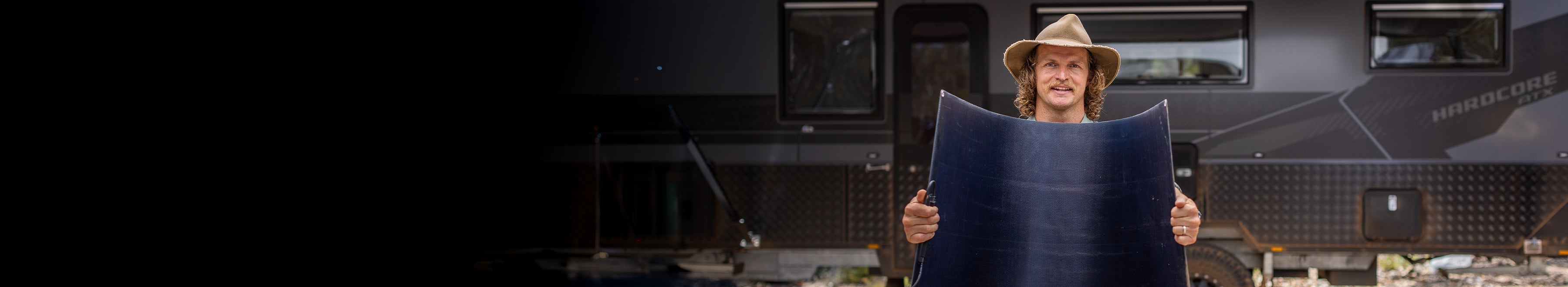 A person in a wide-brimmed hat holds a large flexible solar panel in front of a dark-colored RV. The background is partially in shadow.