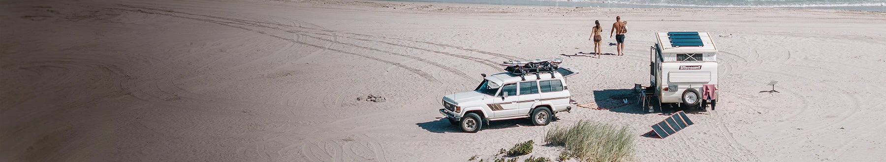 A white SUV with surfboards on top and a camper trailer are parked on a sandy beach. Two people are standing near the shore, with tire tracks visible in the sand.