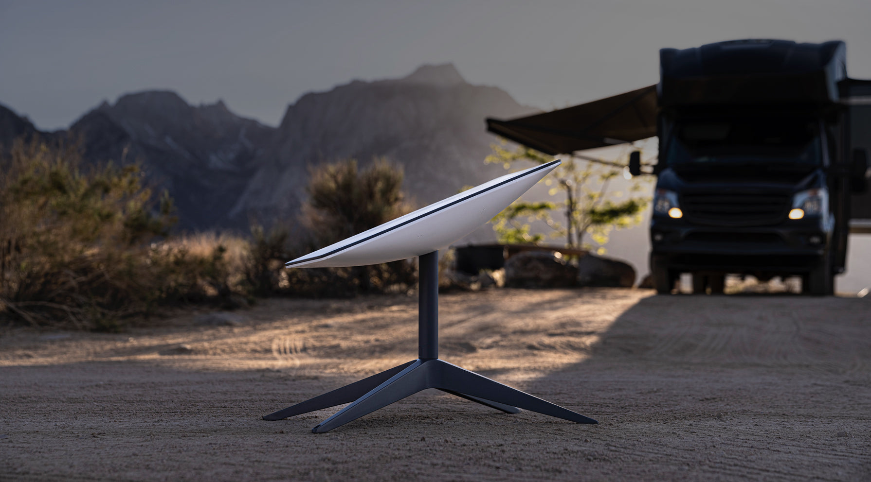 A Starlink satellite dish stands on sandy ground outdoors with mountains in the background and a dark RV parked nearby, partially shaded by an awning.