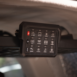 A close-up of the iTechworld 12 GANG Wireless Switch Panel installed in a vehicle, showing labeled buttons for 4WD gear like air compressor, fridge, lights, stereo, hot water, and more.