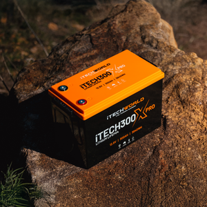 An iTechworld iTECH300X PRO Deep Cycle Lithium Battery (LiFePO4 300Ah, Bluetooth) in black and orange rests on a sunlit rock outdoors, showcasing its specs and branding—ideal for off-grid power needs.