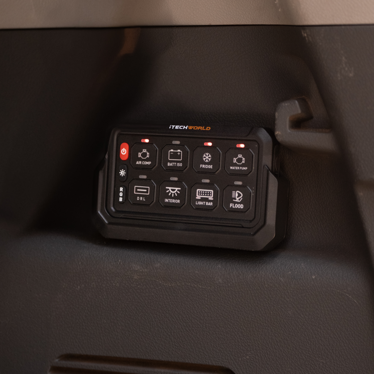 An iTechworld 8 Gang Wireless Switch Panel is mounted on a vehicle dashboard, with labeled buttons for air compressor, lights, fridge, water pump, and other 12V accessories; some indicator lights are illuminated.