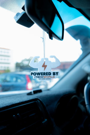 A car rearview mirror displays an iTechworld | Air Freshener | New Car Scent, shaped like flexing arms with a lightning bolt and Powered by iTechworld text, inside a vehicle with a blue-tinted windshield.