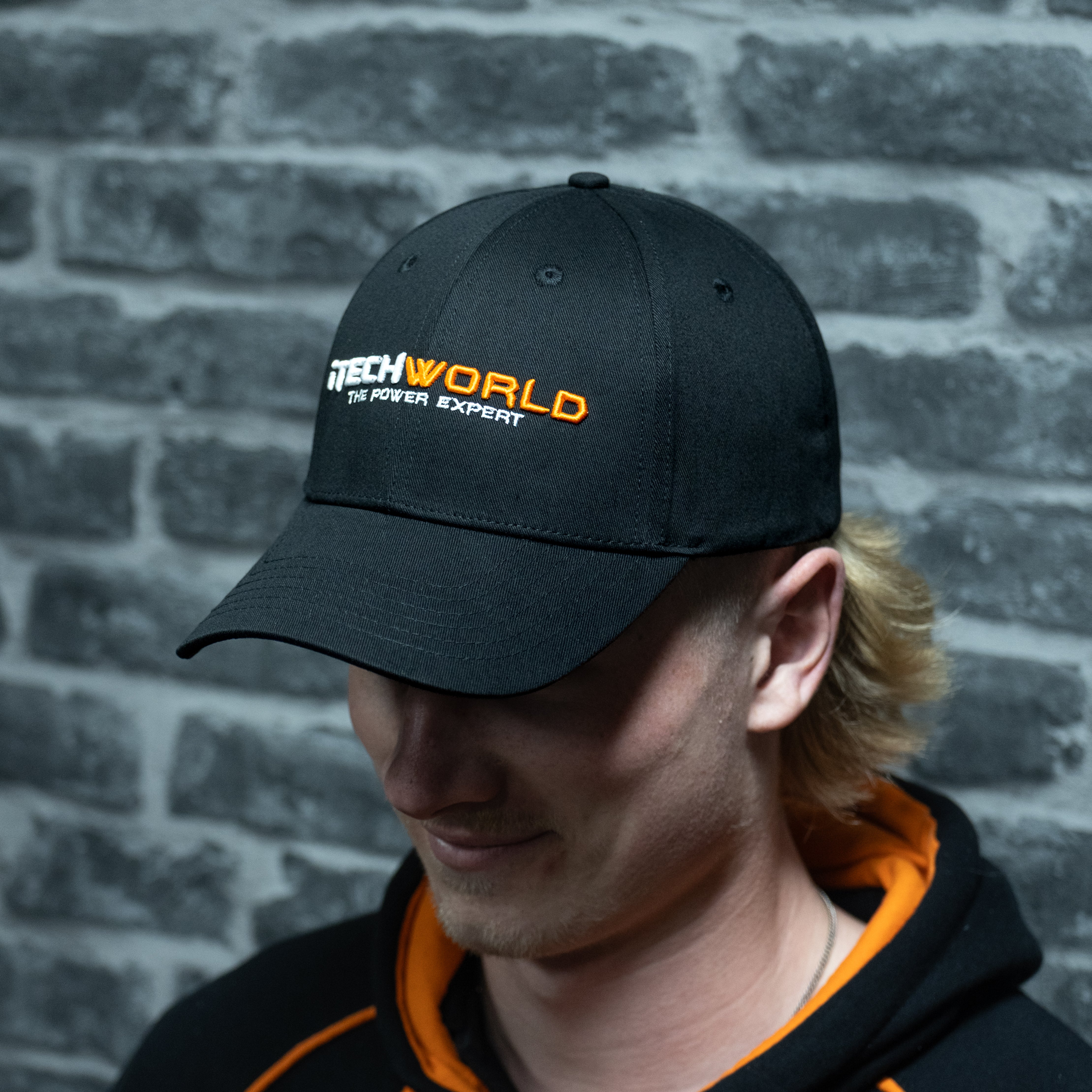 A person in an iTechworld embroidered hat with an adjustable strap and breathable cotton stands by a gray brick wall, pairing it with a black hoodie featuring orange lining.