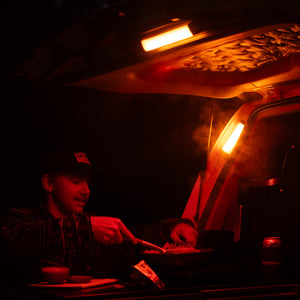 At night, a person sits at the back of a car with the trunk open, illuminated by the iTechworld Magnetic LED Strip Light (300LM), serving food onto a steaming plate with a beverage can nearby.