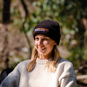 A smiling woman wears the iTechworld | Beanie | Embroidered and a light sweater while relaxing outdoors, sunlight on her face. The beanie is the perfect cold-weather essential for enjoying crisp days against a leafy background.