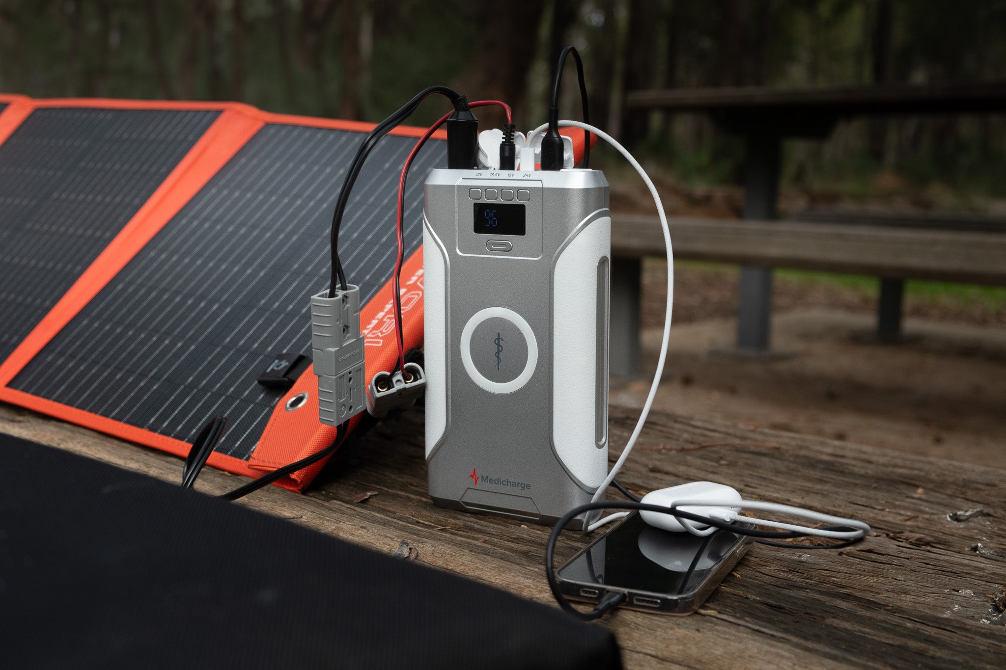 A Medicharge Flex | CPAP Battery | 276Wh sits on a wooden picnic table, powering a CPAP machine, smartphone, and solar panel—an ideal portable power supply for off-grid outdoor adventures.