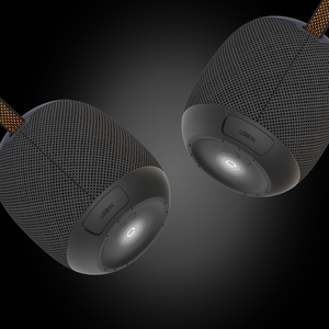 Two iTechworld Pulse Mini waterproof portable speakers, black and cylindrical with mesh exteriors, hang against a dark gradient backdrop. Each features a USB-IN port, power button at the base, and a fabric cord for easy portability.