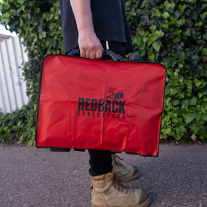 Someone holds the iTechworld Redback | Inverter Generator Protective Cover RB2 in bright red, outdoors among greenery and a metal fence, showcasing its durable protection for generators.