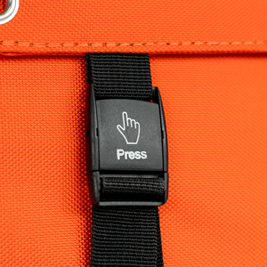 Close-up of a black plastic buckle labeled Press with hand icon on a black strap, attached to bright orange fabric—part of the 400W Solar Blanket with Raptor Skin—with a metal grommet visible in the corner.