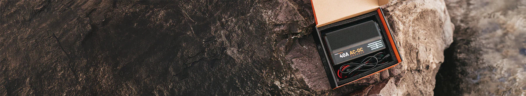 A black electronic device with cables sits inside an open box placed on rocky terrain, partially slid out from behind a large rock.