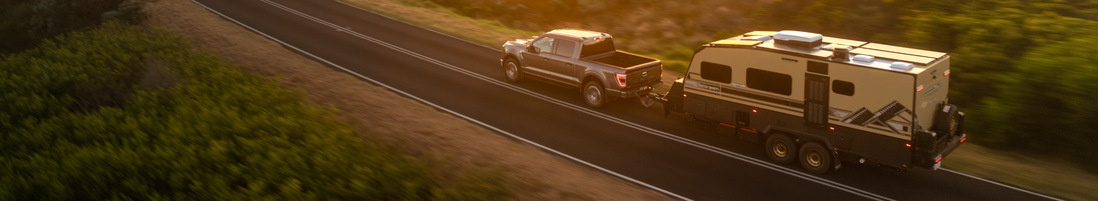 A white off-road vehicle tows a large, modern camper trailer with solar panels, parked on sandy terrain beside a blue coastline and sparse vegetation.