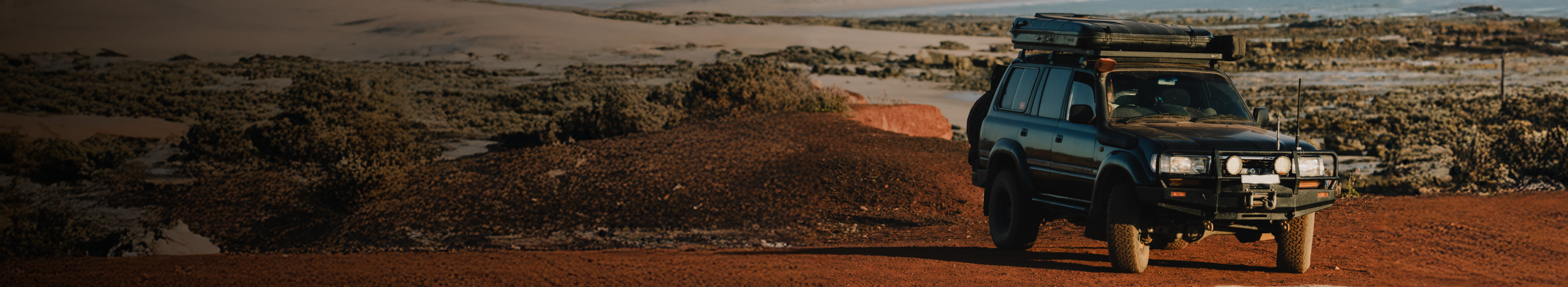 A rugged off-road vehicle with rooftop gear drives on a dirt path through a remote, arid landscape with shrubs and distant sandy hills under a hazy sky.
