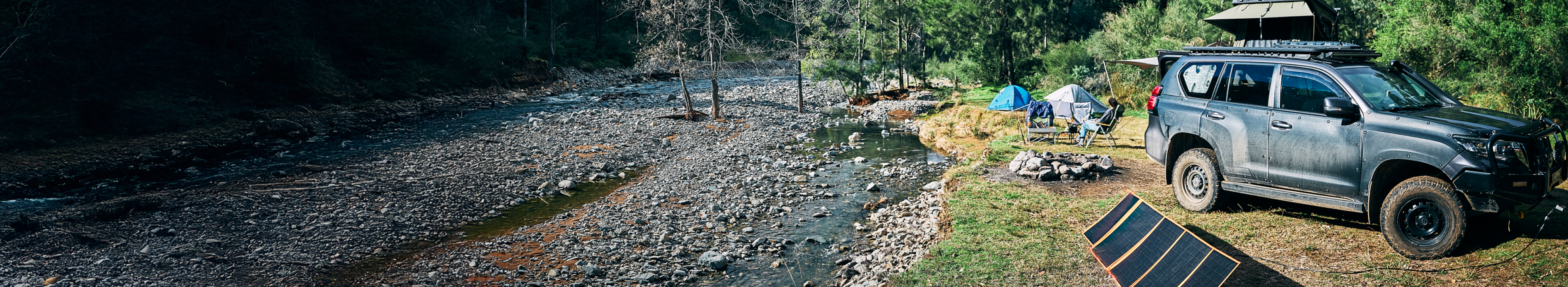 A campsite beside a rocky river, featuring a parked SUV with camping gear, a rooftop tent, chairs, a campfire ring, and a solar panel set up on the grass surrounded by trees.