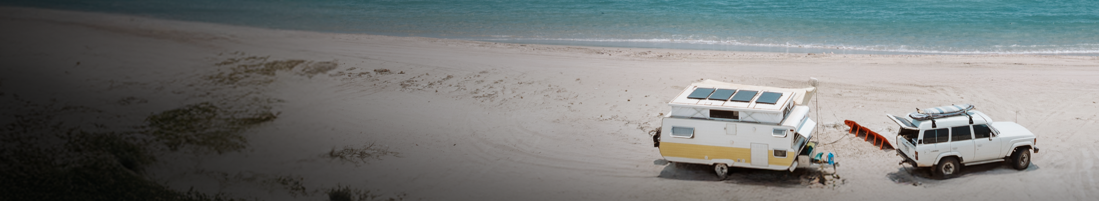 A white SUV towing a yellow and white camper trailer is parked on a sandy beach near the ocean, with a kayak and outdoor gear set up beside it. The sea is calm and blue in the background.