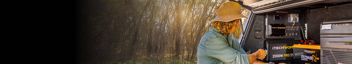 A person wearing a wide-brimmed hat uses power equipment set up in the back of a vehicle in a wooded outdoor area with sunlight filtering through the trees.
