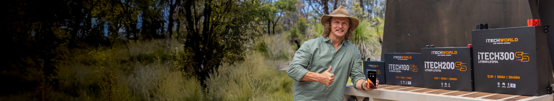 A man in a hat stands outdoors, smiling and giving a thumbs up next to several black lithium battery units displayed on a counter, with trees and greenery in the background.