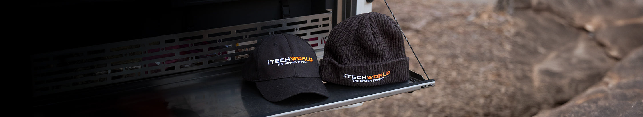 A black baseball cap and a black beanie, both with the TECHWORLD logo, rest on a metal surface outdoors next to some rocks.