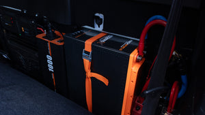A close-up of two iTechworld iTECH100SX Slim Deep Cycle Lithium Batteries (Bluetooth LifePO4 100Ah), black and orange, secured with an orange strap and connected by thick red/blue cables, installed near a black carpeted vehicle floor.