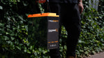 A person in dark clothing holds an iTechworld iTECH100SX Slim Deep Cycle Lithium Battery with Bluetooth LifePO4 100Ah, featuring an orange handle, while standing outdoors against a leafy green background.