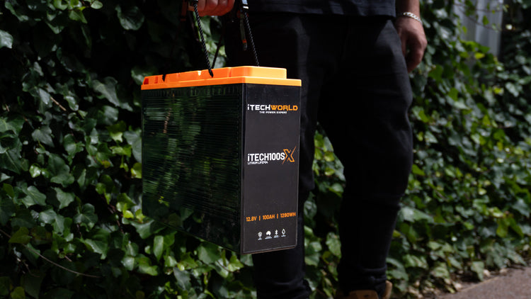 A person in dark clothing holds an iTechworld iTECH100SX Slim Deep Cycle Lithium Battery with Bluetooth LifePO4 100Ah, featuring an orange handle, while standing outdoors against a leafy green background.