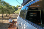 A man sits on a mat beside a white camper van in a grassy, tree-lined area, petting a brown dog. The van’s back is open, revealing an iTechworld 2000W Pure Sine Wave Inverter (12V to 240V) neatly installed inside—perfect for powering essentials on any caravan adventure.