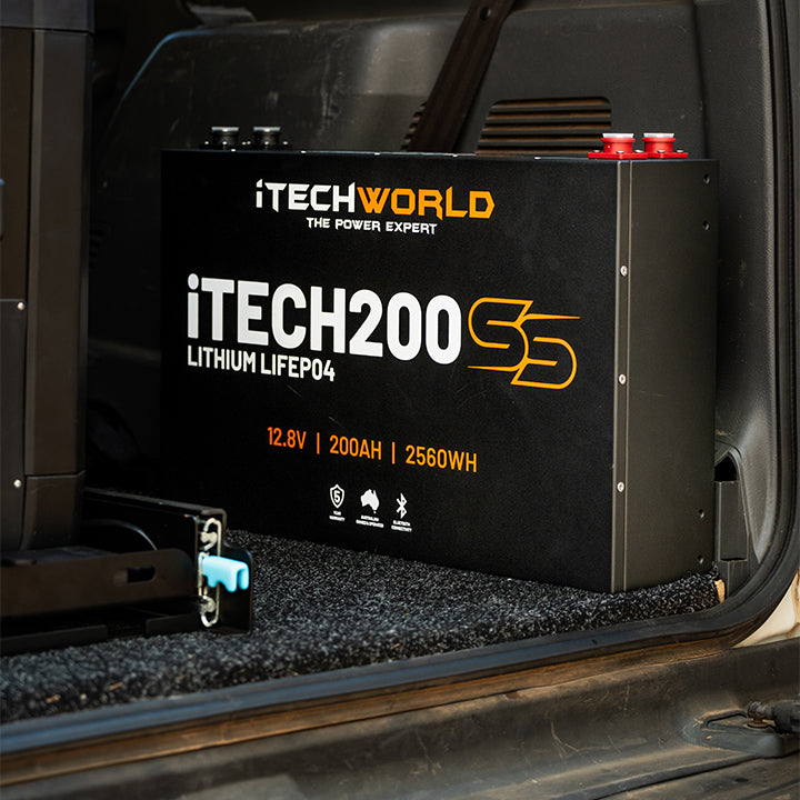 A large iTechworld iTECH200SS Super Slim Deep Cycle Lithium Battery (12.8V, 200Ah, 2560Wh, LiFePO4 with Bluetooth) featuring orange and white text is installed in the back of a vehicle on a carpeted surface.