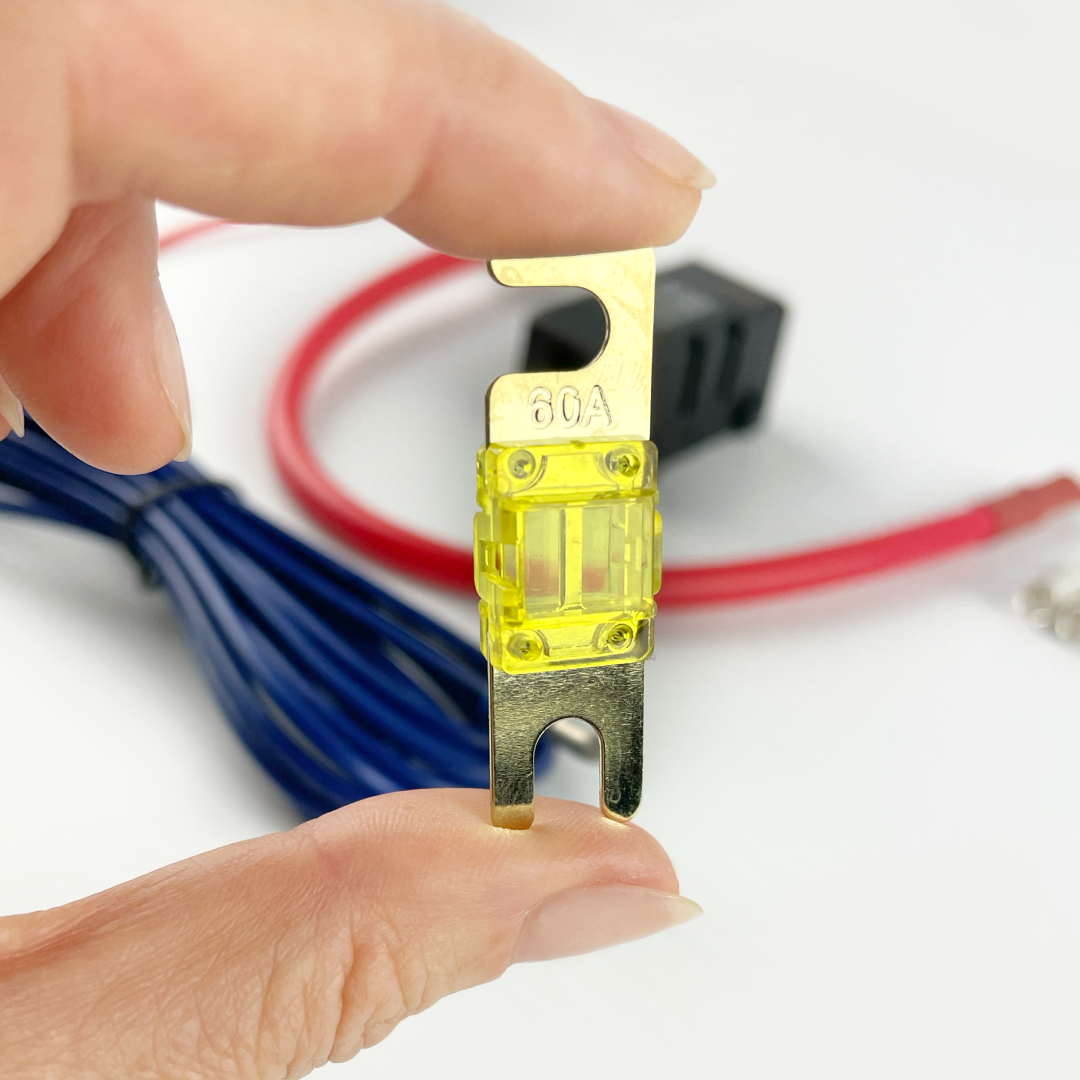 Close-up of a hand holding a yellow 60A automotive fuse, with blue wire and red 6AWG cable from the iTechworld Plug and Play 40A DCDC Wiring Kit blurred in the background, shown as part of a dual battery system setup.