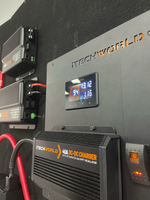 Close-up of the iTechworld iTECHBM500 Battery Monitor with Shunt (500A) and display panel showing 13.12V and 1.16A, providing precise battery readings. Red and black cables are connected, alongside an amp-hour counter and additional charger units mounted on a black wall.