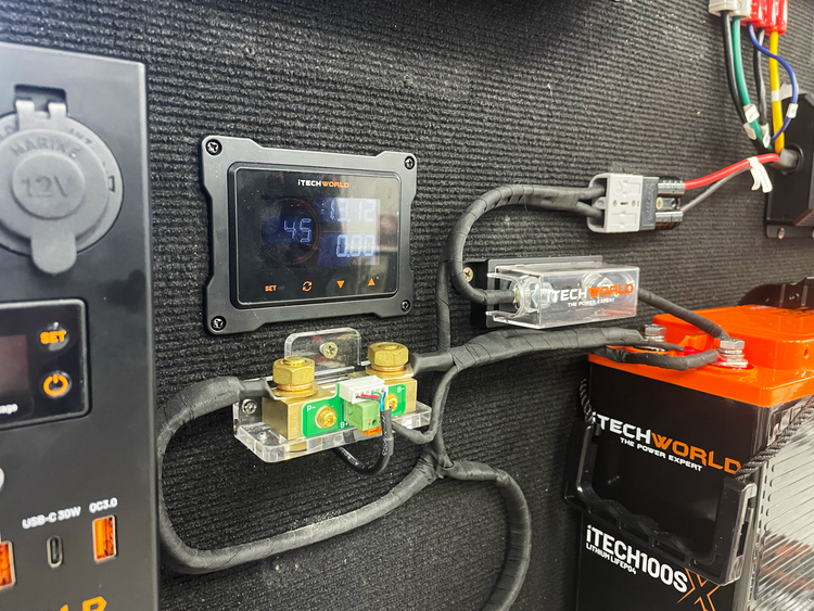Close-up of an electrical setup featuring a digital display showing voltage and amperage, fuses, cables, a USB port, and a red iTechworld battery pack, all mounted on a textured black panel. The integrated iTECHBM500 Battery Monitor with Shunt by iTechworld enables precise amp-hour counting and measurement with its 500A shunt.