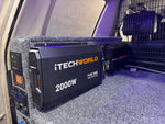 An iTechworld 2000W Pure Sine Wave Inverter (12V to 240V) is securely installed in a vehicle, mounted on carpeted flooring with nearby electrical components and a metal safety barrier visible in the background. This dependable inverter provides efficient power conversion for mobile and off-grid use.