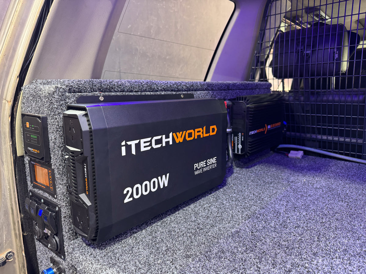An iTechworld 2000W Pure Sine Wave Inverter (12V to 240V) is securely installed in a vehicle, mounted on carpeted flooring with nearby electrical components and a metal safety barrier visible in the background. This dependable inverter provides efficient power conversion for mobile and off-grid use.