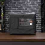An iTechworld portable power station is positioned on a wooden table in front of a gray brick wall and black metal shelves, safeguarded by the iTechworld Power Station Cover PS2000 for enhanced durability.