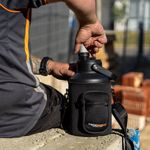 A person in a gray shirt with orange accents opens an iTechworld | 3.8L Water Bottle, a black insulated jug, while sitting on concrete at a construction site with bricks and fencing in the background.