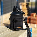 The iTechworld | 3.8L Water Bottle, a large black vacuum-sealed bottle with handle and zippered pocket, sits outdoors on concrete with blurred construction materials in the background. The iTechworld brand is printed on its leak-proof cover.