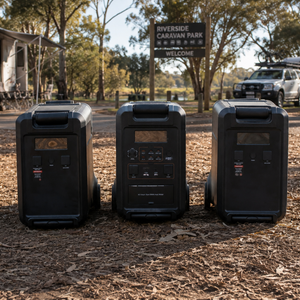 Three iTechworld PS3600 Portable Lithium Power Stations with EB3840 Expansion Battery Kits are placed outdoors near trees, a car, and a Riverside Caravan Park sign—ideal for home backup or camping adventures.