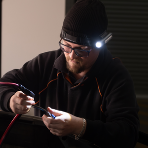 A man in glasses and a beanie works intently in a dim room, illuminated only by the ultra-bright 450LM beam of his iTechworld Magnetic Head Torch as he connects wires, dressed in a dark sweatshirt and focused on his task.