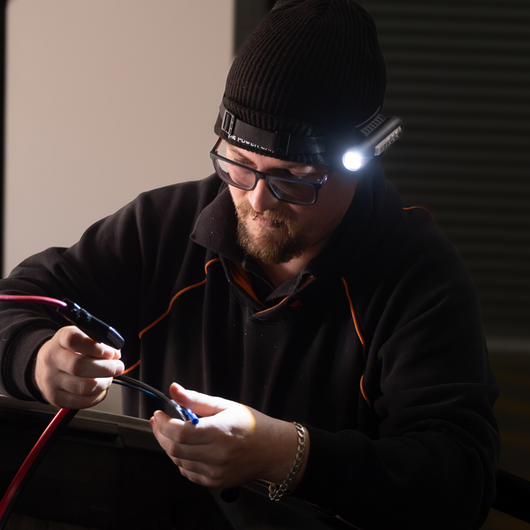 A man in glasses and a beanie works intently in a dim room, illuminated only by the ultra-bright 450LM beam of his iTechworld Magnetic Head Torch as he connects wires, dressed in a dark sweatshirt and focused on his task.