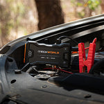 An iTechworld JS60 Lithium Jump Starter and Backup Power Bank with Heavy-Duty Case, featuring red jumper cables, is connected to a car battery under the hood in a wooded outdoor setting.