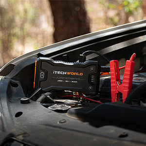 An iTechworld JS60 Lithium Jump Starter and Backup Power Bank with Heavy-Duty Case, featuring red jumper cables, is connected to a car battery under the hood in a wooded outdoor setting.
