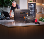 An iTechworld Off-grid backup kit (PS2000 + JS80) with red clamps sits on a kitchen counter, while a man works in the background.