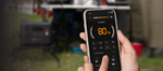 Someone holds a smartphone showing 80% battery on the iTechworld Off-grid Backup Kit (PS2000 + JS80) app, with a portable generator and outdoor gear visible in the blurred background.